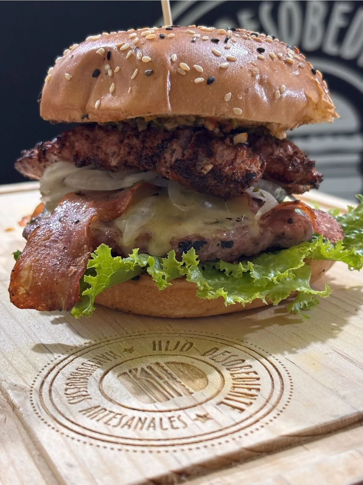Hamburguesa con tocino, queso y lechuga sobre la tabla de madera con el sello de El Hijo Desobediente